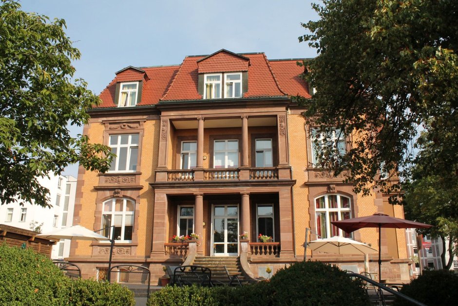 Klinik MKK Talitha – Blick auf die Vorderseite mit Terrasse und Grünflächen Frontalansicht der MKK Talitha mit Bäumen und Sonnenschirmen im Vordergrund